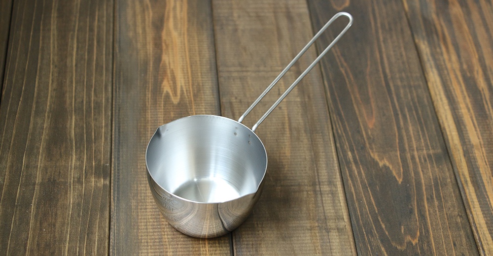 不鏽鋼長柄量杯，帶有一個細長的單柄，杯身為圓形，表面呈現銀色金屬光澤。