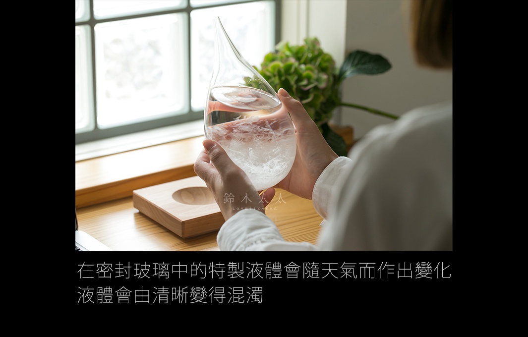 一隻手正捧著透明的玻璃製天氣瓶，瓶內有類似結晶的白色沉澱物，瓶身呈水滴狀，頂端細長。玻璃瓶被放置在一個淺木色的木製底座上。