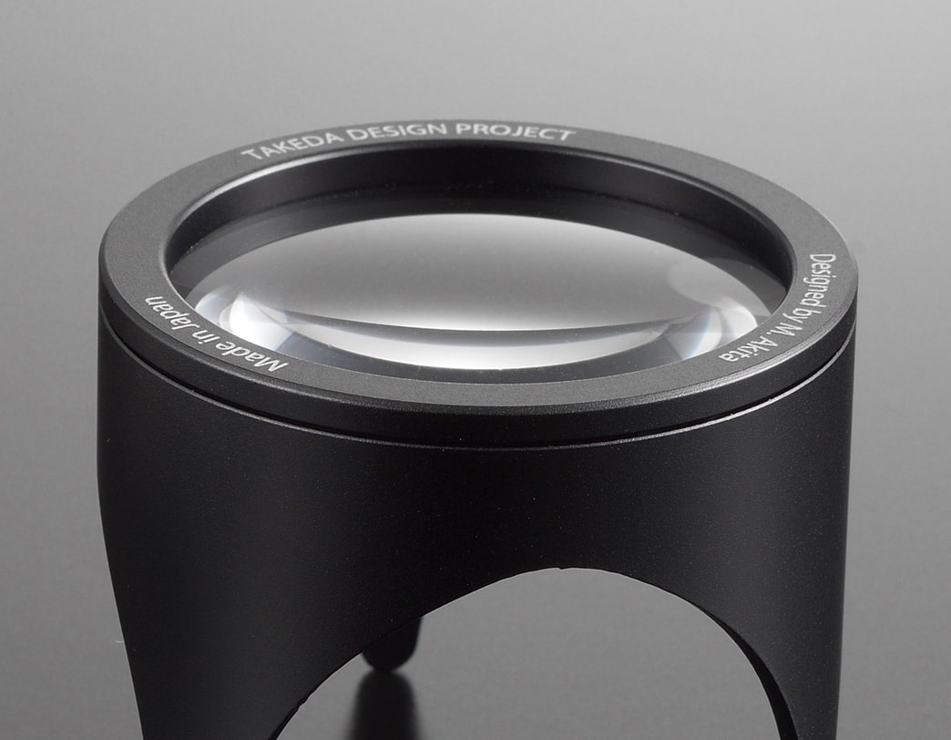 黑色鋁製閱讀放大鏡，鏡圈刻有「TAKEDA DESIGN PROJECT」、「Made in Japan」、「Designed by M. Akira」字樣，放大鏡鏡片透明。
