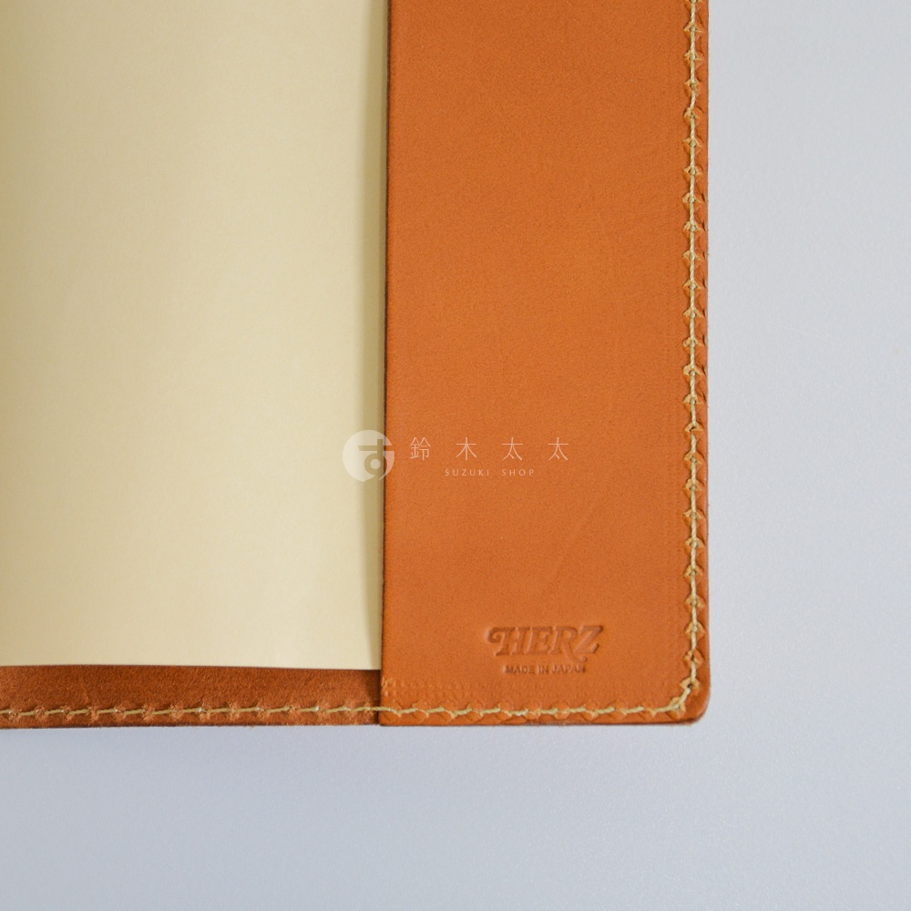 這是一張近距離拍攝的日本HERZ職人手作皮革書套特寫，展示了溫潤的焦糖色厚皮革質感，邊緣處有精緻的米白色手工縫線。皮革右下角壓印有HERZ與MADE IN JAPAN字樣，圖片中央並有鈴木太太SUZUKI SHOP的水印標誌。