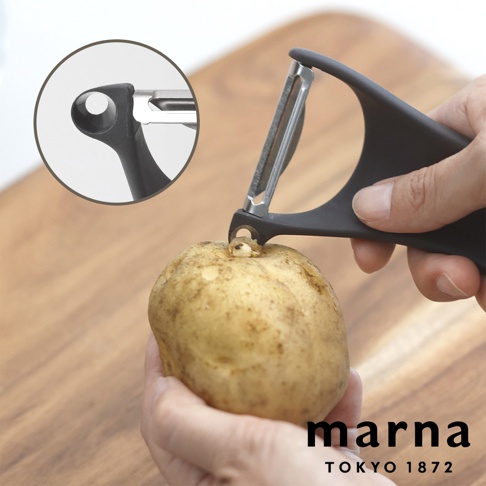 一隻手正使用 Marna 品牌的黑色多功能削皮器挖除馬鈴薯的芽眼，削皮器前端設計有專用的挖芽尖端，左上角圓形放大視窗特寫了該挖芽孔結構，背景為木紋砧板。