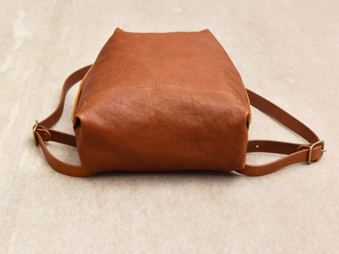 職人手作皮革後背包，顏色為棕褐色，材質為真皮，包身呈現簡約褶皺風格，配有可調節的皮革肩帶及黃銅色金屬扣環。