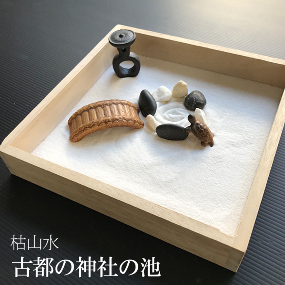 木製淺盤中鋪著白色細砂，擺放著日式枯山水模型，包含一座棕色木質拱橋、黑色和白色鵝卵石、一個黑色裝飾物，以及一隻棕色小烏龜，砂上 rake 出波浪紋。