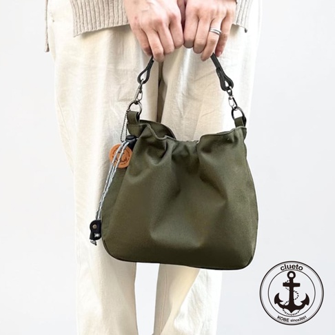 一位女性雙手提著一個神戶clueto品牌的深橄欖綠色BALON mini防潑水手提包，包身設計簡約，採用柔軟布料製成並帶有皺褶質感，側面懸掛著帶有品牌標誌的圓形木質吊牌與繩索細節，右下角印有clueto KOBE的錨形商標。