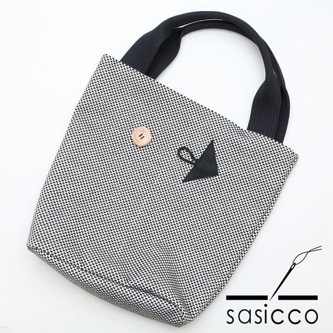 三河木綿刺子織紋樣的直式收納托特包，黑白格紋織紋，配有黑色織帶提把，正面有一個木質鈕扣及一個黑色三角形裝飾。