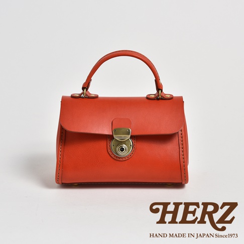 職人手作皮革迷你手提包，採用橘紅色皮革，配有黃銅色金屬鎖扣和手提把，側邊有縫線細節。