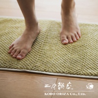 一雙赤腳踩在淡黃色棉麻混紡人字紋浴墊上，浴墊邊緣為白色滾邊，背景為木質地板。