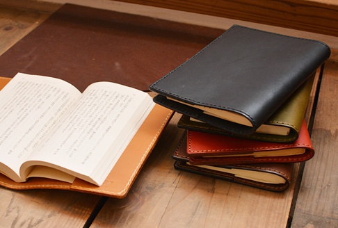 職人手作皮革厚革書套，展示黑色、橄欖綠、紅棕色與深棕色等多種顏色。左側為一本攤開的書，置於淺棕色皮革書套內，書套邊緣有細緻的縫線。右側堆疊著四個不同顏色的皮革書套，頂部為黑色。