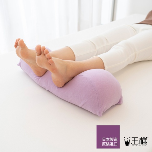 淺紫色半月形抬腿枕，表面布料柔軟，放置於白色床鋪上，一位女性的雙腳舒適地倚靠在枕頭上，腳踝和腳底部分清晰可見。