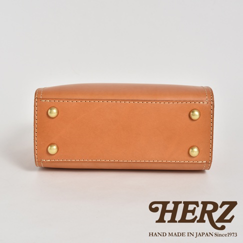 職人手作皮革兩用方形單肩包，以天然皮革製成，呈現溫潤的焦糖棕色，包身底部設有四顆黃銅鉚釘，搭配細緻的縫線，展現精湛工藝。