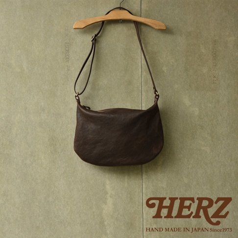 職人手作皮革Organ單肩半月包，深棕色，光滑皮革材質，半月形輪廓，單肩背帶，拉鍊開合，掛於木質衣架上。