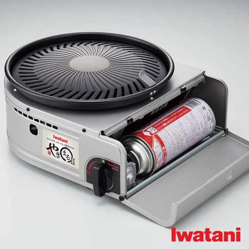 Iwatani CB-SLG-2 無煙燒烤爐，銀色金屬機身，搭配黑色圓形烤盤。烤盤表面有放射狀紋路，爐體側面設有瓦斯罐槽，可放入紅色瓦斯罐。