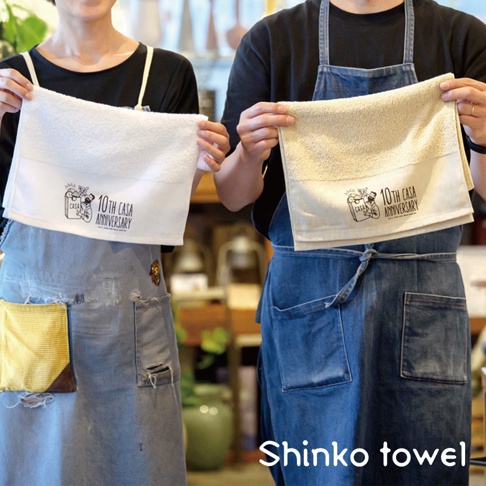 兩位身穿圍裙的人手中各拿一條素面毛巾，白色毛巾左側印有「CASA 10TH ANNIVERSARY」圖案，米色毛巾右側亦有相同圖案。