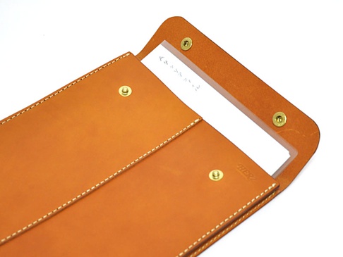 職人手作皮革信封式雙釦手拿袋，採用天然皮革材質，呈現溫潤的棕褐色，車縫線清晰，設有兩個金屬釦及一個透明夾層，可放置文件或卡片。