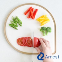 淚滴型白色抗菌料理砧板，邊緣有淺棕色框架。砧板上擺放著切塊的紅甜椒、黃甜椒、蘆筍、花椰菜和切片的番茄，其中一個番茄正被刀切開。