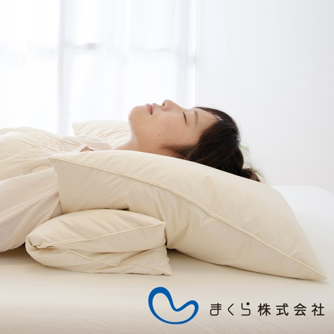 一組兩入的米白色羽絨枕，疊放在床上，一位女子側臥於枕上，畫面背景為明亮的窗簾。