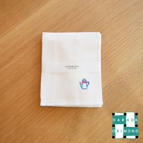 白色八重紗精繡擦拭巾，織有網格紋理，左下方繡有藍色與紅色線條點綴的茶壺圖案，產品標籤為白色，印有日文及英文產品名稱。