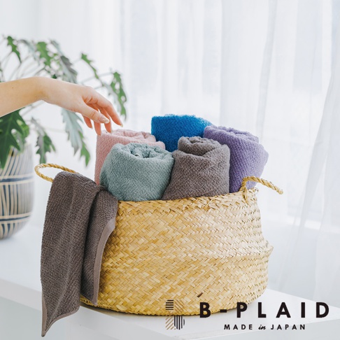 編織籃裡擺放著捲起的 EVE 今治浴巾，顏色包含粉色、藍色、紫色、灰色和綠色。其中一條棕灰色浴巾掛在籃子外側。