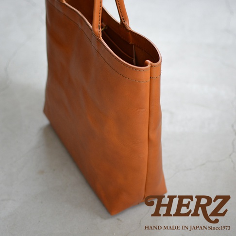 HERZ品牌標誌，下方有HAND MADE IN JAPAN Since1973字樣，圖片主體為一個棕色皮革托特包，包身線條簡潔，手柄部分略有彎曲，整體呈現職人手工製作的質感。