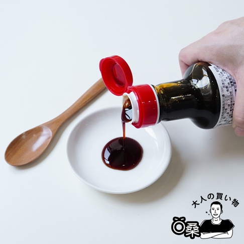 一瓶深褐色醬汁從紅色瓶蓋的瓶子中倒入白色小碟中，旁邊放著一個木湯匙。