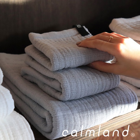 堆疊的灰色無撚紗毛巾，表面有紋理，一隻手觸摸著最上方的毛巾，手腕上戴著戒指。