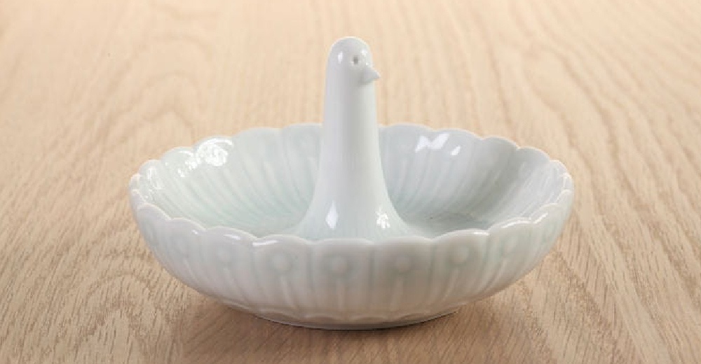 白色陶瓷製的白鳥造型碟，碟身為扇形，外緣有花瓣狀的紋飾，中央立有一隻向上伸展的鳥形支架，碟內有細膩的放射狀紋路。