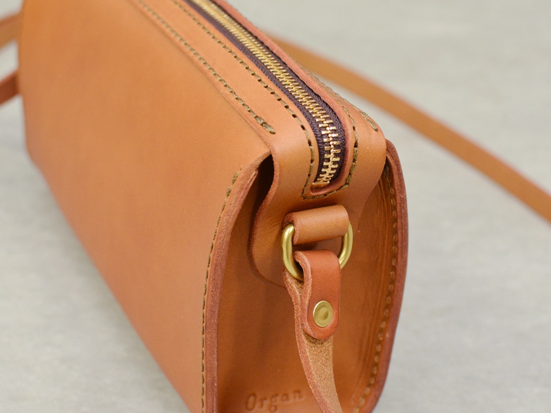 淺棕色皮革製成的手作拉鍊單肩包，具有精緻的縫線和金屬拉鍊，側面有皮革繫帶固定，風格簡約。
