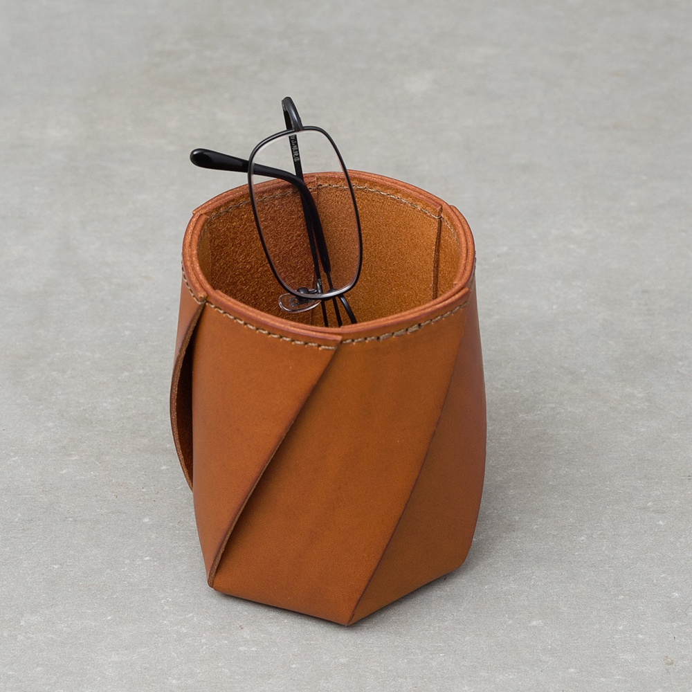 職人手作皮革Organ六角收納筆筒，採用棕色皮革製成，筆筒側邊呈現獨特的摺疊設計，上方放入一副黑色細框眼鏡。