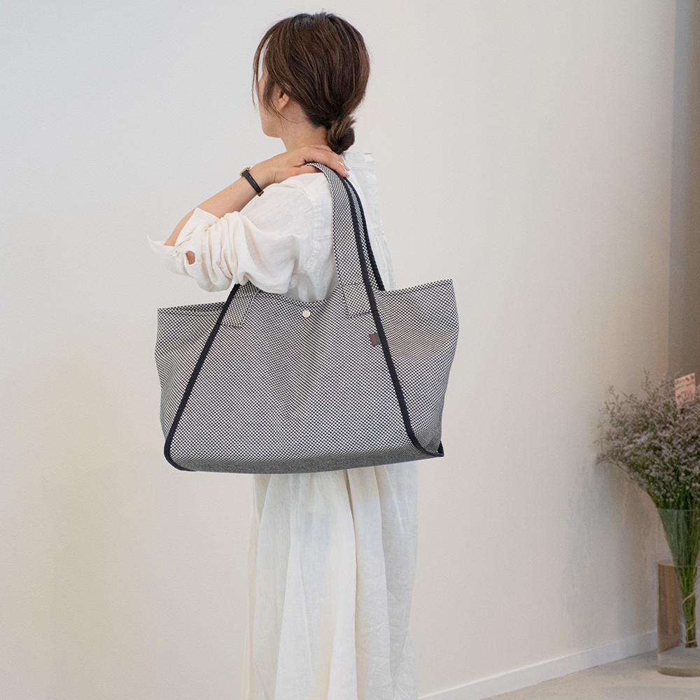 一位女性背著一個大容量的刺子織托特包，包身為白色底黑色細點圖案，配有黑色織帶提把。