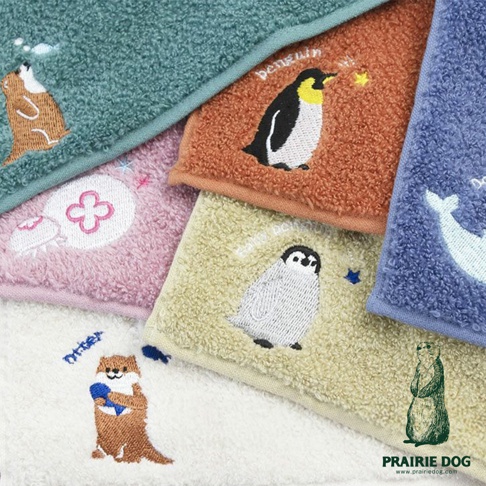 多條毛巾以不同顏色和動物刺繡呈現，包括綠色毛巾上的海獺，橘色毛巾上的企鵝與星星，粉色毛巾上的花朵圖案，淺黃色毛巾上的企鵝，以及藍色毛巾上的鯨魚。白色毛巾上有一隻拿著球的海獺。