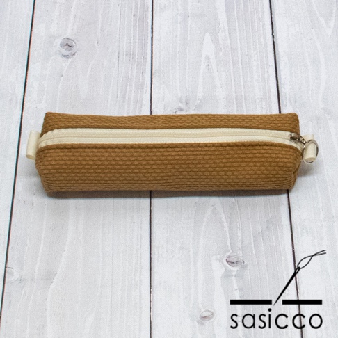 棕色三河木綿刺子織長條筆袋，配有米白色拉鍊和銀色拉鍊頭，袋口兩側有小掛環。