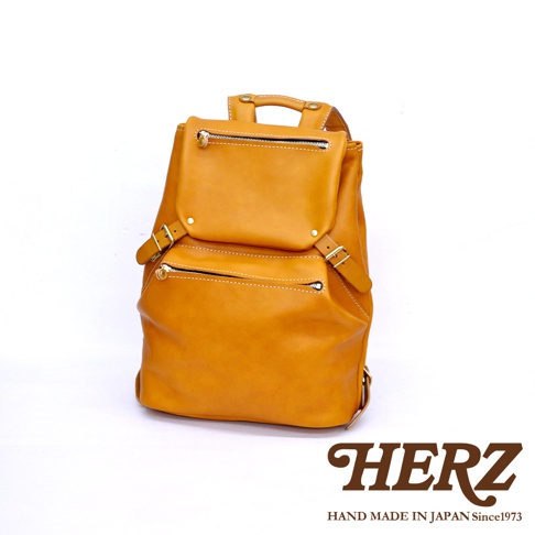 一個黃褐色皮革後背包，有頂部提把、兩個前置拉鍊口袋，以及側邊的扣帶和調整環。