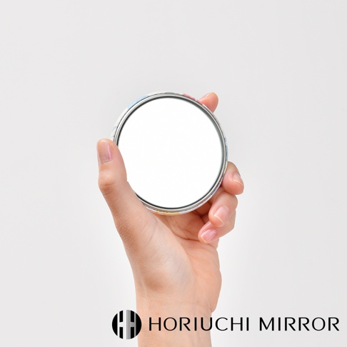 一隻手拿著一個圓形口袋小鏡子，鏡面為白色，外框有彩色圖案，鏡子邊緣為銀色金屬材質。