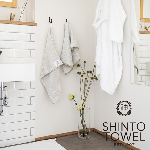 浴室角落，兩個淺米色紋理毛巾掛在牆上，左側毛巾有品牌標籤。右側掛著一件白色浴袍。一束乾燥花草插在透明玻璃花瓶中，旁邊有兩朵淺黃色小花。牆面為米白色，左側是白色長方形磁磚牆面。