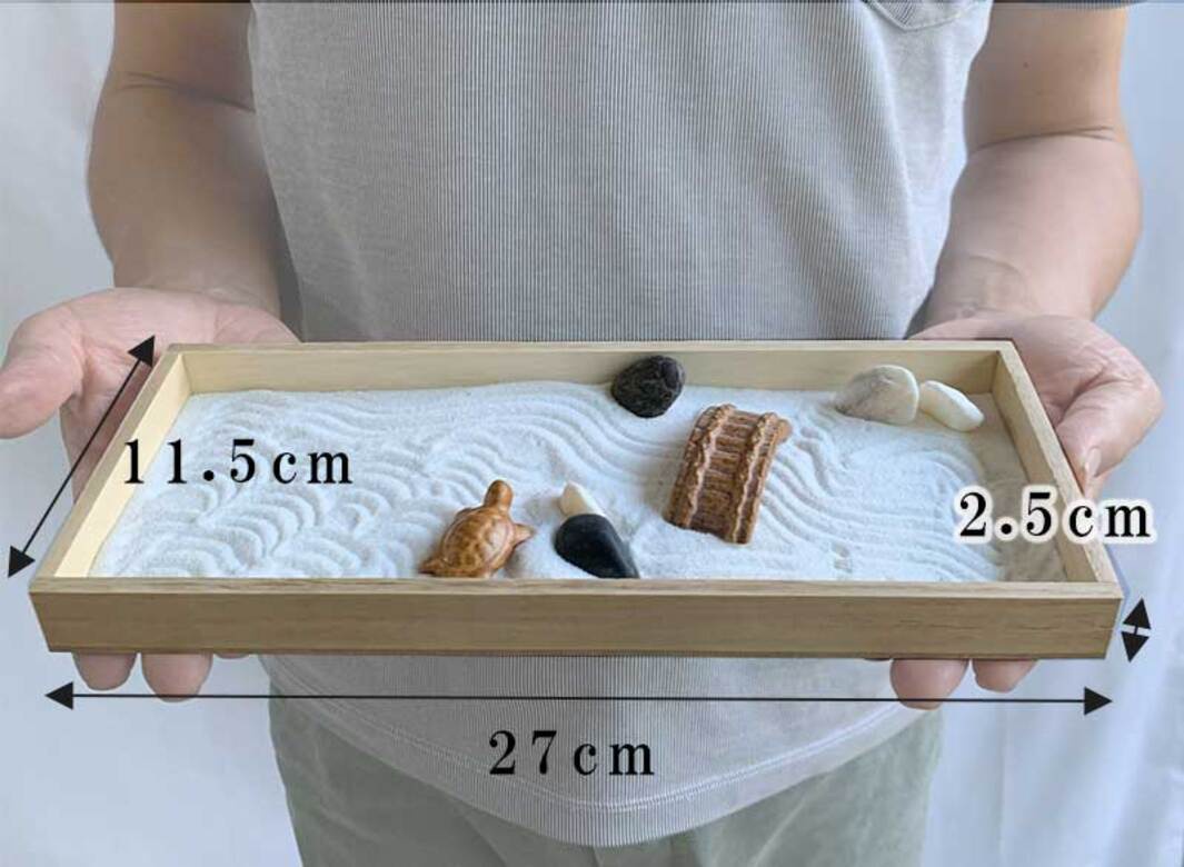 木製淺盤內盛有白色細沙，沙上堆出波浪紋理。盤中有一個淺棕色陶瓷烏龜擺飾、一個黑色尖角擺飾、一座褐色拱橋模型、一顆深色卵石及兩顆白色卵石。尺寸標示為長 27 公分，寬 11.5 公分，高 2.5 公分。