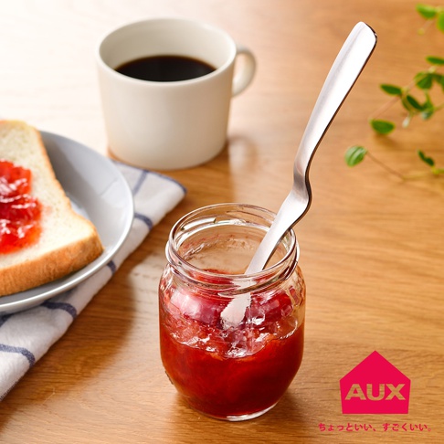 銀色金屬製的不沾桌果醬抹刀，插入裝有紅色果醬的透明玻璃罐中，一旁有塗抹果醬的吐司和一杯咖啡。