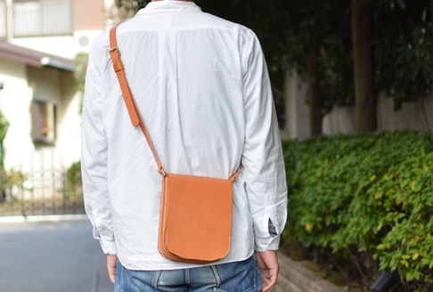 棕色皮革翻蓋直式單肩包，配有可調節的同色系皮革背帶，斜挎於身穿白色長袖襯衫的人身上。