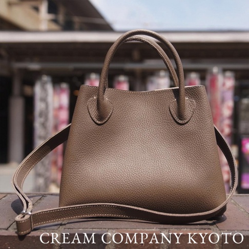 深棕色粒面皮革手提包，附有可拆卸肩帶，包包上有縫線細節，背景為模糊的建築物。