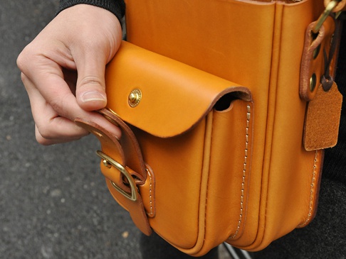 職人手作皮革皮扣直式單肩包，採用天然鞣製皮革，呈現溫潤的焦糖色。包身結構清晰，縫線紮實，配有金屬皮扣及可調節的皮革背帶，展現簡約實用的設計風格。