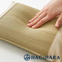 手壓在藺草編織的枕頭上，展示其柔軟的質地。枕頭表面為天然藺草材質，色澤呈現米白色與淺棕色交織，邊緣有淺米色的布料包覆。