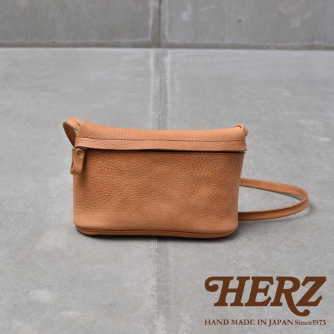 HERZ 品牌標誌，下方有 HAND MADE IN JAPAN Since1973 字樣，搭配淺棕色牛皮製成的貼身斜背包，包包呈現長方形，表面有細緻紋理，上方有拉鍊開合，右側有細長背帶。