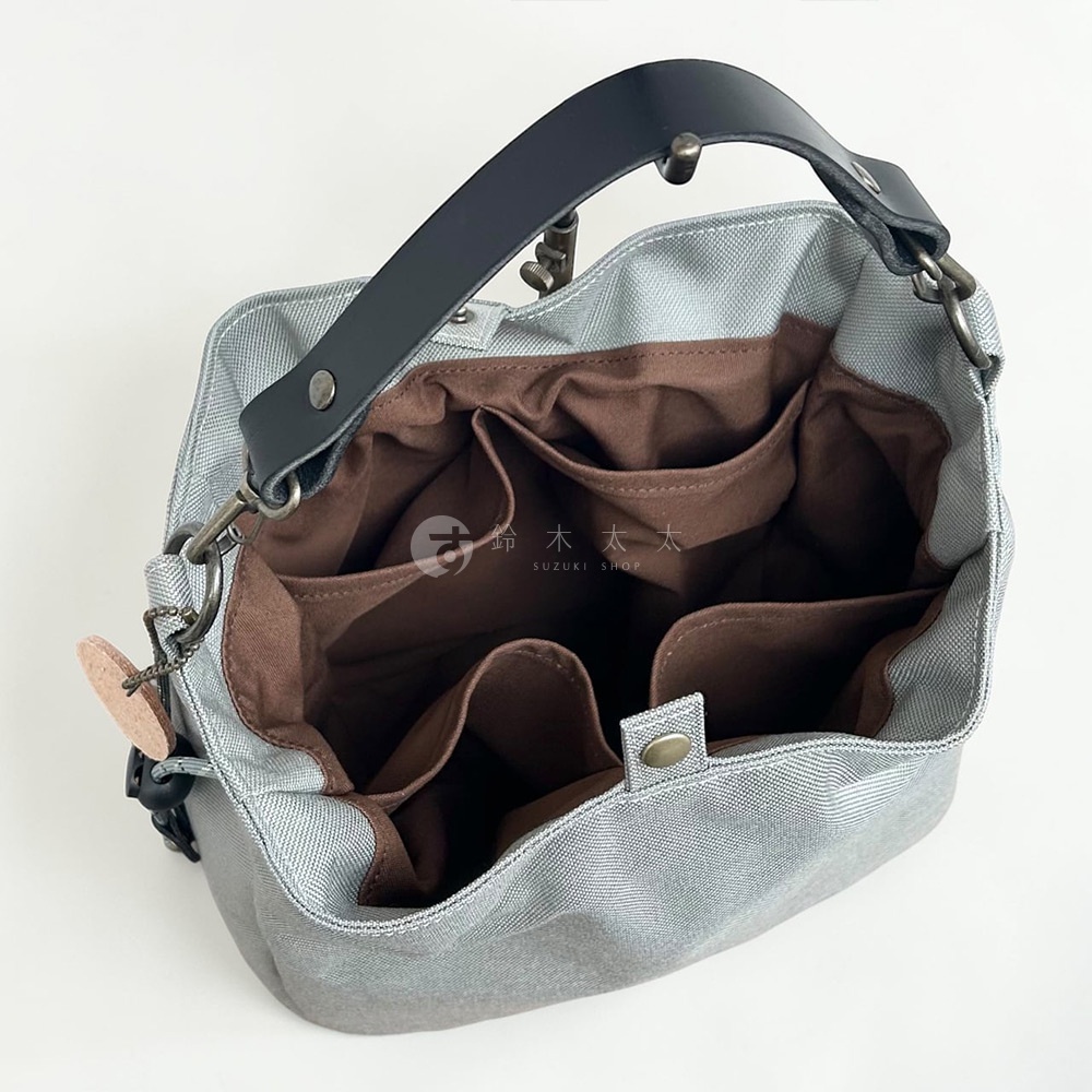 一張神戶clueto品牌的BALON mini防潑水手提包俯視圖，包身呈現灰色織物質感，內部配有深褐色布料隔層與多個收納口袋。包包上方設有一條黑色皮革提把，並配有金屬釦具與一個圓形皮革吊飾，袋口中央則有一處金屬按扣設計。