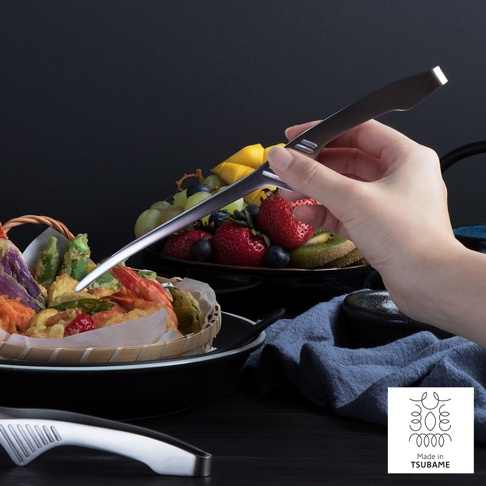 手持銀色不鏽鋼公筷夾，夾取籃中炸物。背景為一盤色彩鮮豔的水果，包含草莓、奇異果、葡萄和芒果。