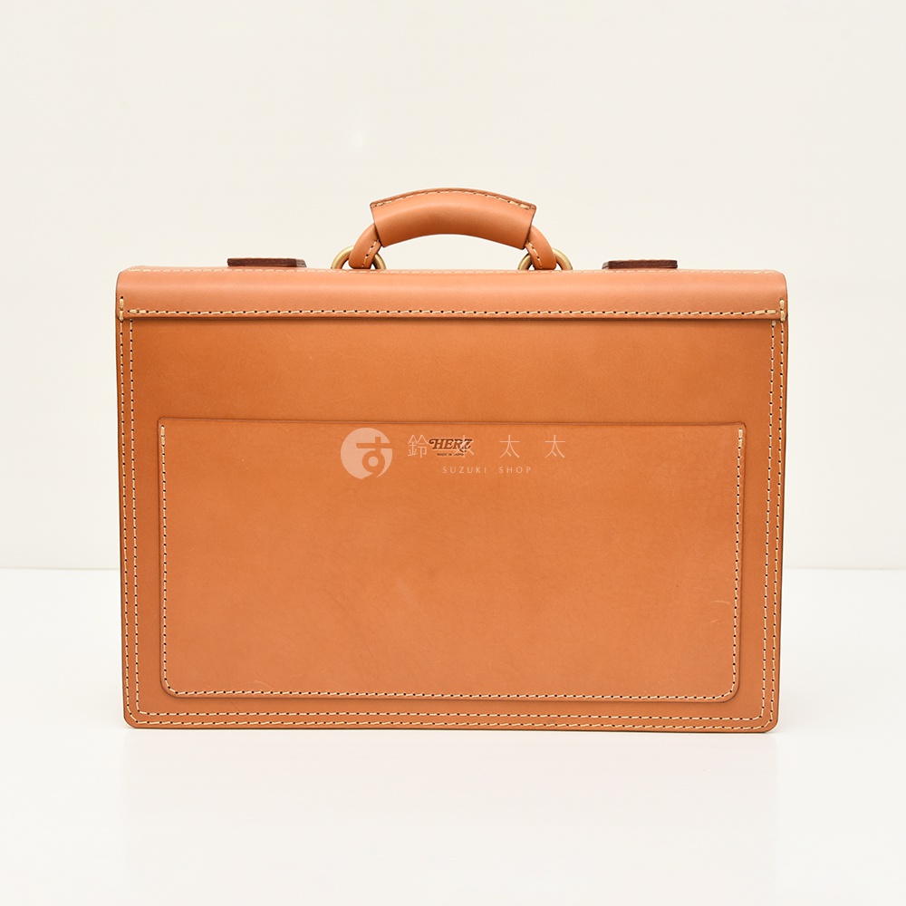 職人手作皮革機師箱，採用高品質天然皮革，呈現溫潤的淺棕色調。箱體設計簡潔，頂部設有皮革提把，兩側有金屬扣環，正面有一個大面積的貼袋，整體以細緻的縫線勾勒出質感。