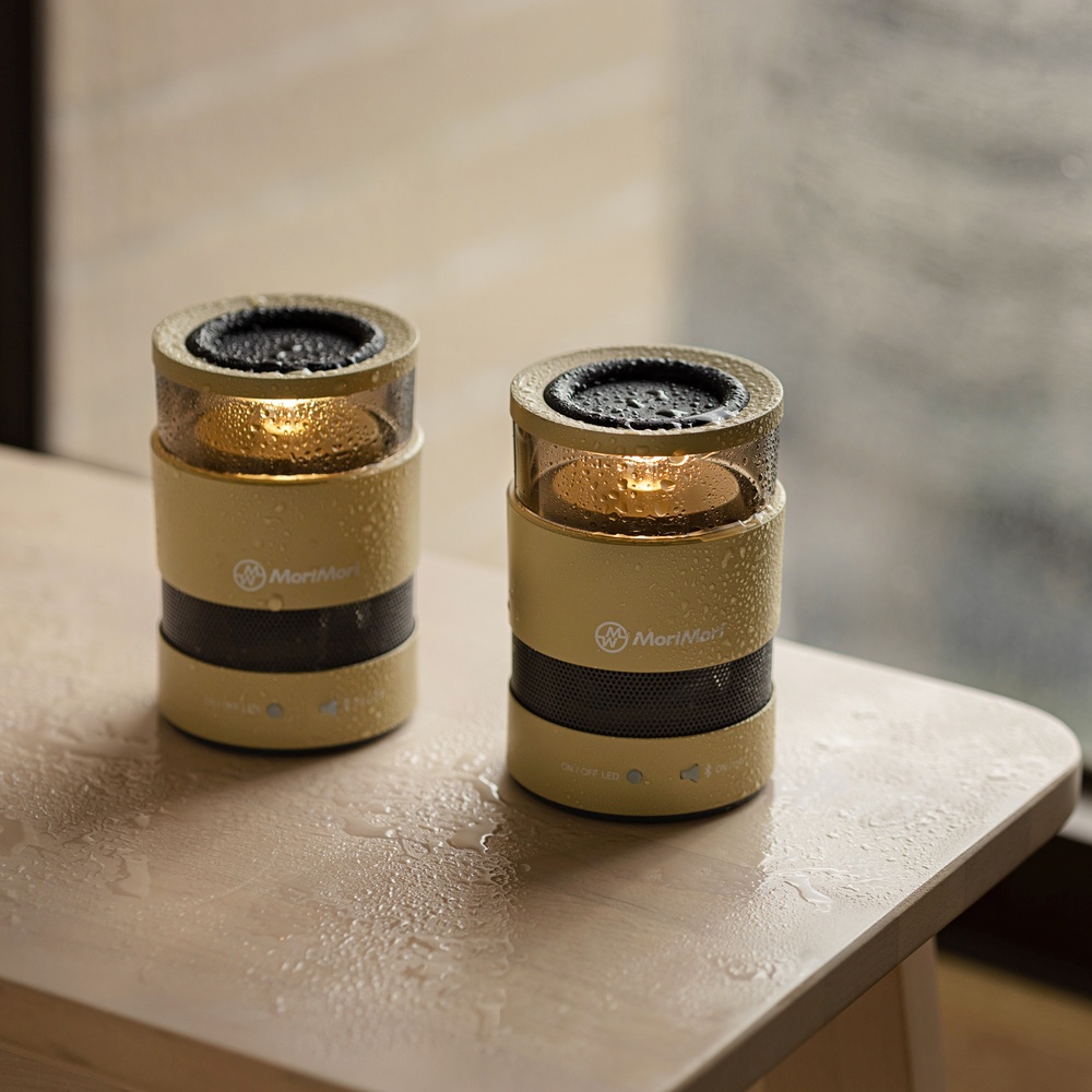 兩台圓柱形戶外無線藍牙氛圍燈音響，皆為淺黃色，頂部為黑色網狀喇叭，中間為透明燈罩，內含暖黃色燈光，表面佈有水珠。