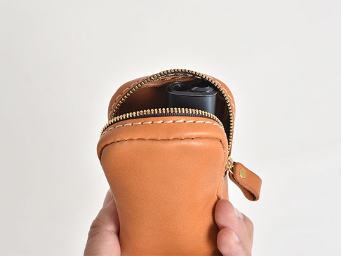 手持棕色皮革製成的手工菸盒收納包，拉鍊部分打開，露出內部一個黑色的小型電子產品。