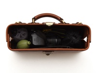 職人手作皮革橫長形杜勒斯單肩包 (CW-4)，內裝有黑色相機、綠色吹氣球、灰色和黑色收納袋，以及 Nikon 相機背帶。包身為棕色皮革材質，配有金色金屬鎖扣和提把。