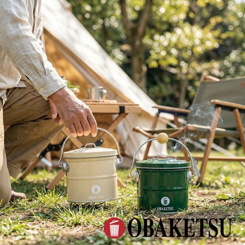 模式B： 戶外場景中，一個人彎腰準備拿起一個米白色的OBAKETSU蚊香桶，旁邊還有一個綠色的同款蚊香桶，兩者都有金屬提把，其中綠色桶蓋冒著些許煙霧，背景是帳篷和露營椅。