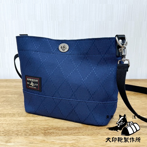 深藍色刺子織迷你側背包，織有菱形紋路，設有銀色金屬轉鎖和黑色織帶肩帶。包包左側有一個帶有日文文字的黑色方形品牌標籤。