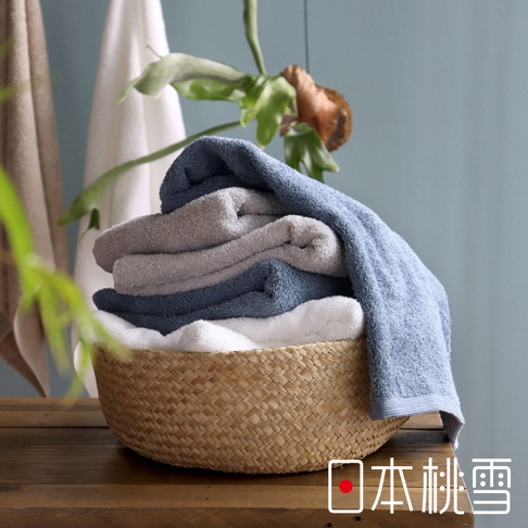 竹編籃中堆疊著藍色、灰色和白色的精梳棉毛巾，背景為淺藍色牆面與綠葉裝飾。